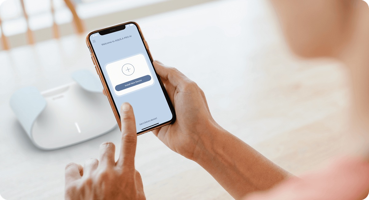 Hands holding a phone showing the AQUILA PRO AI welcome screen and an Install New Device button, with the router sitting on the table behind it.
